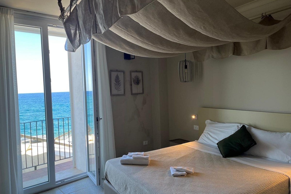 A hotel room with a bed, a view of the sea through a sliding door, and a ceiling fan.