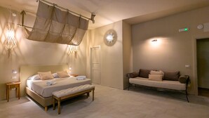 A bedroom with a bed, bench, bedside table, hanging canopy, wall-mounted light, and a sofa. - Palazzo Russo Ortigia Suite & Apartments (Syracuse)