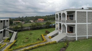 Um edifício de vários andares com fachada de pedra, um pátio central com gramado e canteiros de flores, e vista para a paisagem ao redor.