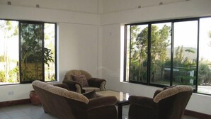 Quarto espaçoso com duas poltronas marrons e uma mesa pequena, janelas amplas com vista para o verde, e piso em azulejos.
