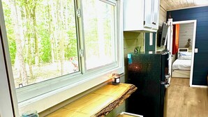 Eine kleine Küche mit einer Holzausziehtafel, zwei weißen Hocker, einem schwarzen Kühlschrank und einem Fenster mit Blick auf einen Wald.