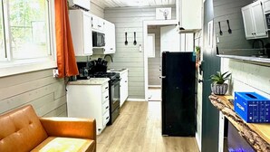 Eine kompakte Küche mit Mikrowelle, Herd und Kühlschrank. Es gibt eine kleine Essecke mit einer Couch und einem Fenster mit Vorhängen.