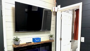Ein Zimmer mit an der weißen Wand montiertem Fernseher, einem eingebauten Kamin mit blau beleuchtetem Feuer und einem Holzregal mit Dekorationsgegenständen.