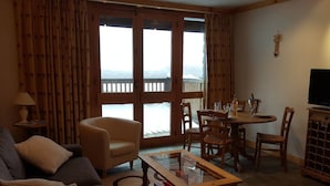 Ein gemütliches Wohnzimmer mit einer Couch, Sesseln und einem Couchtisch mit Glasplatte. Es gibt einen Essbereich mit Tisch und Stühlen sowie einen Fernseher auf einer Holzkommode. Große Fenster mit Vorhängen bieten einen Blick ins Freie.