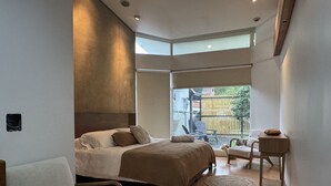 A modern bedroom with a large bed, a sitting area with a chair and a small table, and a view of the outdoors through a large window. - Spacious 5BR in Escazu w/Jacuzzi, Sauna & Fire Pit (San Rafael)