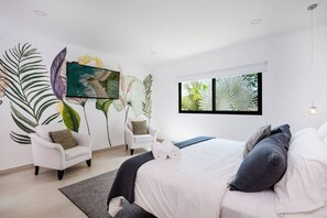 A modern bedroom with a large bed, two white armchairs, a large window with a view of greenery, and a wall mural of tropical leaves. - Luxury, free transfer & Cook, Cinema, Beach Club, (Tamarindo)