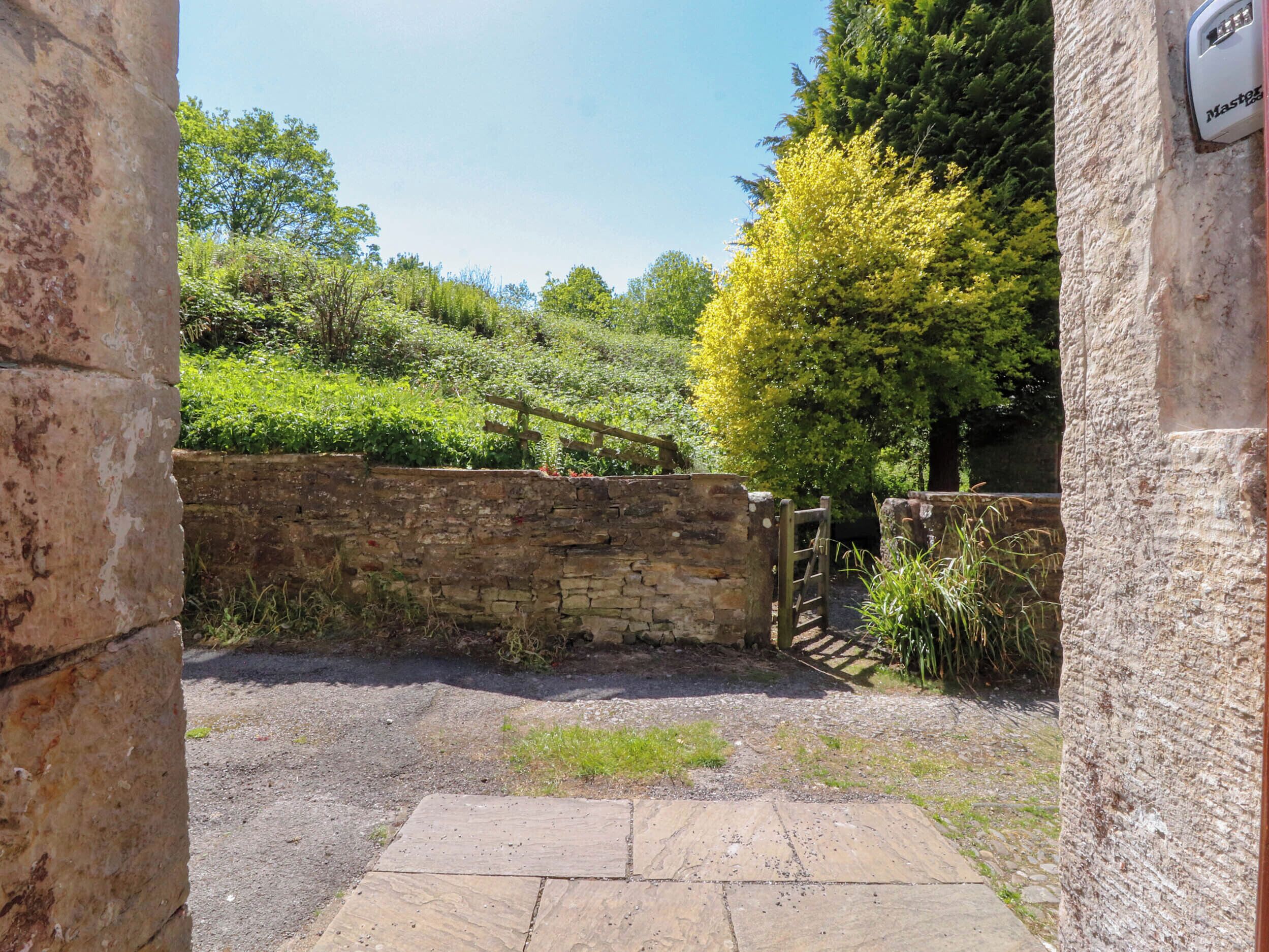 A stone pathway leading to a rustic gate, surrounded by greenery and trees.