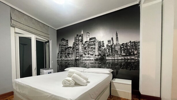 A hotel room with a bed, a nightstand, a window, and a cityscape mural on the wall. - Charming apartment in Castro (Castro-Urdiales)