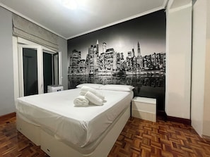 A hotel room with a bed, a nightstand, a window, and a cityscape mural on the wall.