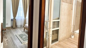 A hotel room with a wooden floor, a green patterned rug, a bed, a bathroom with a glass shower enclosure, and a view of the outside through the window. - Elegant holiday home situated in Salerno, city centre, a few km from Amalficoast (Salerno)