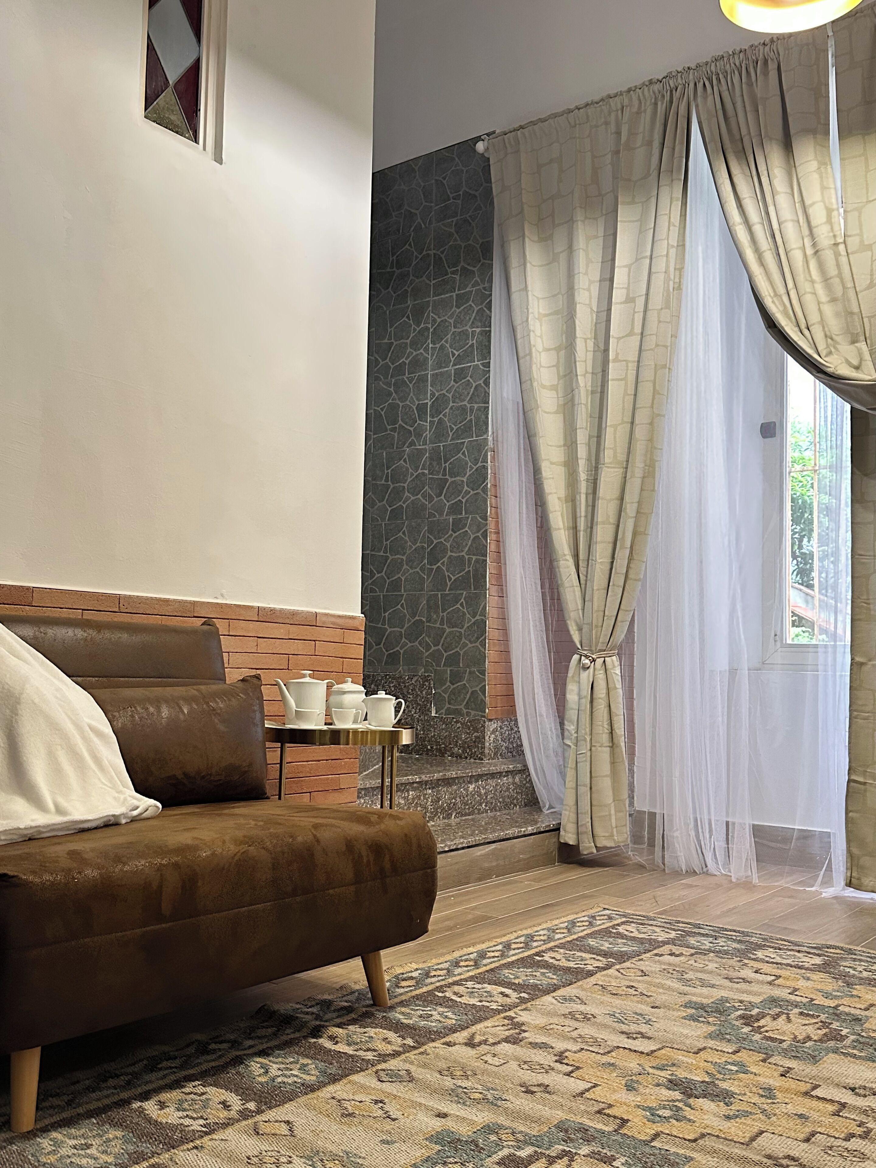 A cozy room with a brown sofa, a small table with tea sets, a patterned rug, a stone accent wall, and sheer curtains.