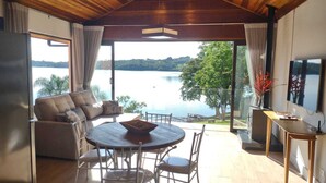 Una sala moderna con un gran ventanal con vistas a un lago, un sofá, una mesa de comedor y un televisor.