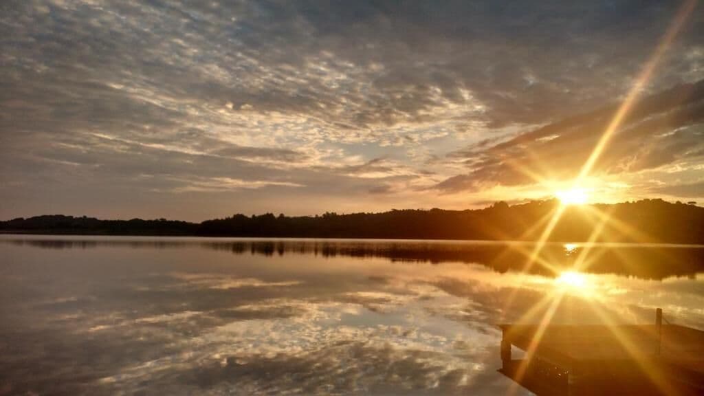 Un lago tranquilo al amanecer, con el sol reflejado en el agua, rodeado por un paisaje montañoso.