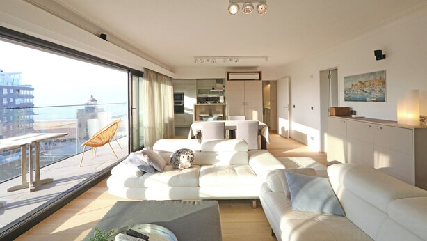 A modern living room with a large sofa, a dining area, and a view of the city. - Apartment in Het Zoute With Sea Views (Knokke-Heist)