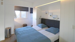 A bedroom with two beds, a window with blinds, a bedside table, a trash can, and a wall-mounted light fixture. - Apartment in Het Zoute With Sea Views (Knokke-Heist)