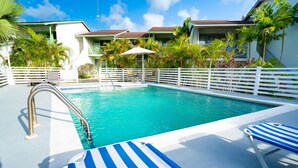 Une piscine entourée d’une clôture blanche, avec des chaises longues à rayures bleues et blanches, et des palmiers.