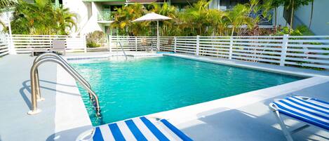 A swimming pool with a white fence, blue and white striped lounge chairs, and palm trees.