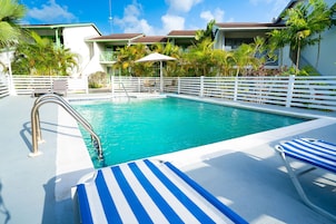 Una piscina con valla blanca, sillas de descanso a rayas azules y blancas, y palmeras.