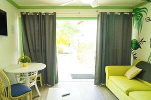 Una habitación con un sofá amarillo, una mesa blanca con dos sillas, un control remoto en el suelo y vistas a un jardín a través de una puerta con cortinas.