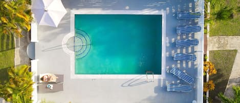 An aerial view of a rectangular swimming pool with a diving board, surrounded by palm trees and lounge chairs.