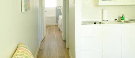 A cozy living area with a patterned sofa, a kitchen with white cabinets, and a hallway leading to a bedroom.
