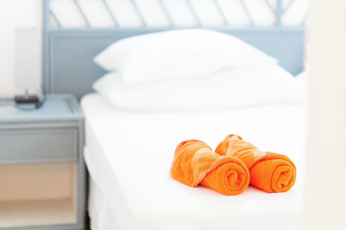 A neatly made bed with white sheets and two rolled orange towels placed on the bed.