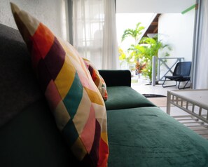 En grön soffa med en färgglad kudde, ett litet bord och en stol i ett ljust rum med utsikt över en trädgård.