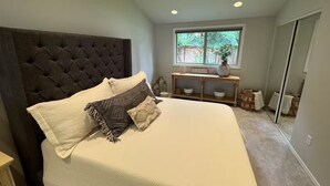 A bedroom with a tufted headboard, a large bed with multiple pillows, a wooden console table with decorative items, and a window with a view of greenery. - Careys Refuge - Family Retreat with Huge Fenced Yard (Bothell)
