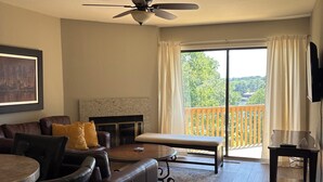 Sala de estar con sofá, mesa de centro y mueble para televisión. Hay un balcón con vistas a árboles y un edificio.