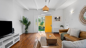A modern living room with a flat-screen TV, a coffee table, a sofa, and a large potted plant. - The Lazy Mimosa Beach Escape Fishing, Bikes & Cozy Firepit (Budgewoi)