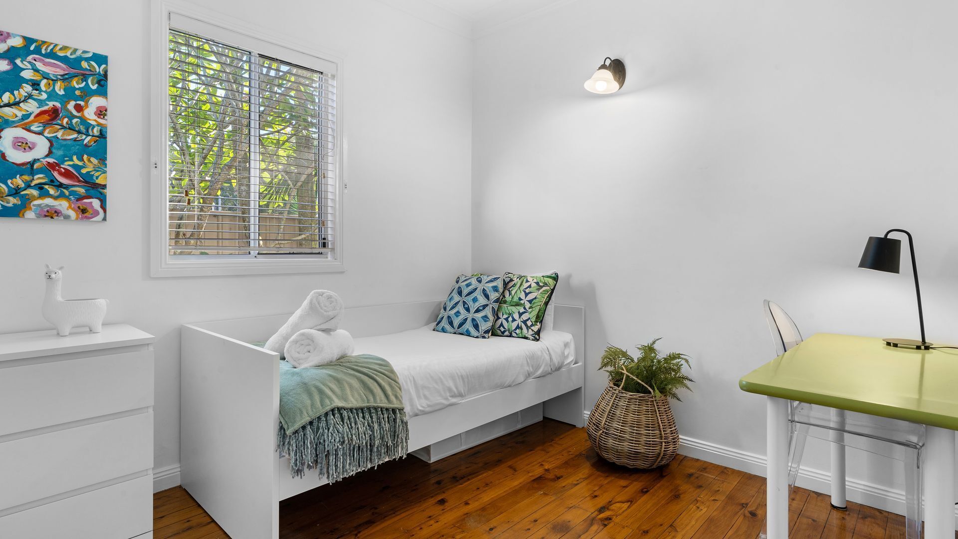 A minimalist bedroom with a single bed, a small desk, a potted plant, and a window with blinds.