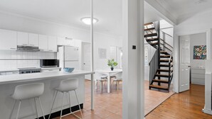 A modern kitchen with white cabinetry, a central island with bar stools, a dining area with a white table and chairs, and a staircase leading to another level. - The Lazy Mimosa Beach Escape Fishing, Bikes & Cozy Firepit (Budgewoi)