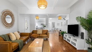 A modern living room with a brown leather sofa, a wooden coffee table, and a TV on a white stand. There is a large mirror on the wall and a hanging light fixture above the sofa. - The Lazy Mimosa Beach Escape Fishing, Bikes & Cozy Firepit (Budgewoi)