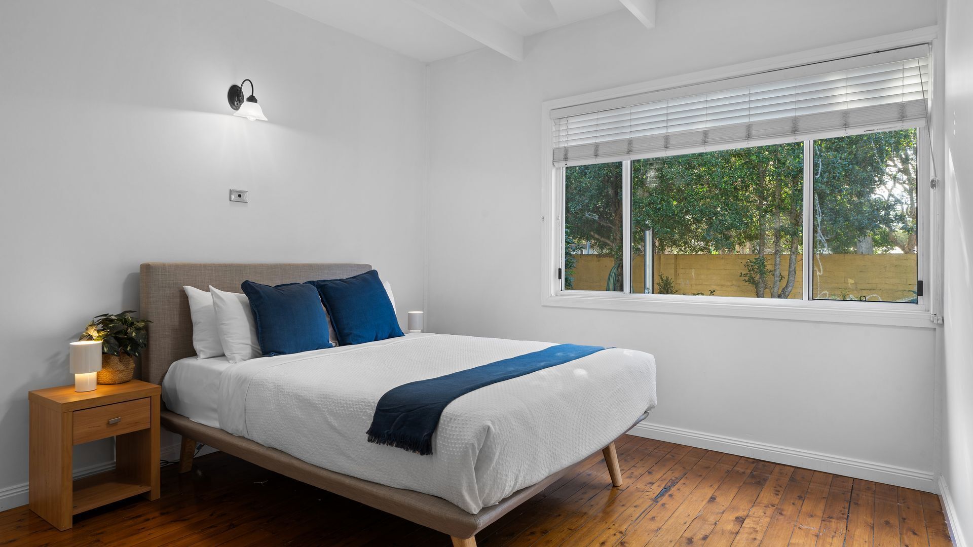 A bedroom with a bed, bedside table, lamp, and a window with blinds.