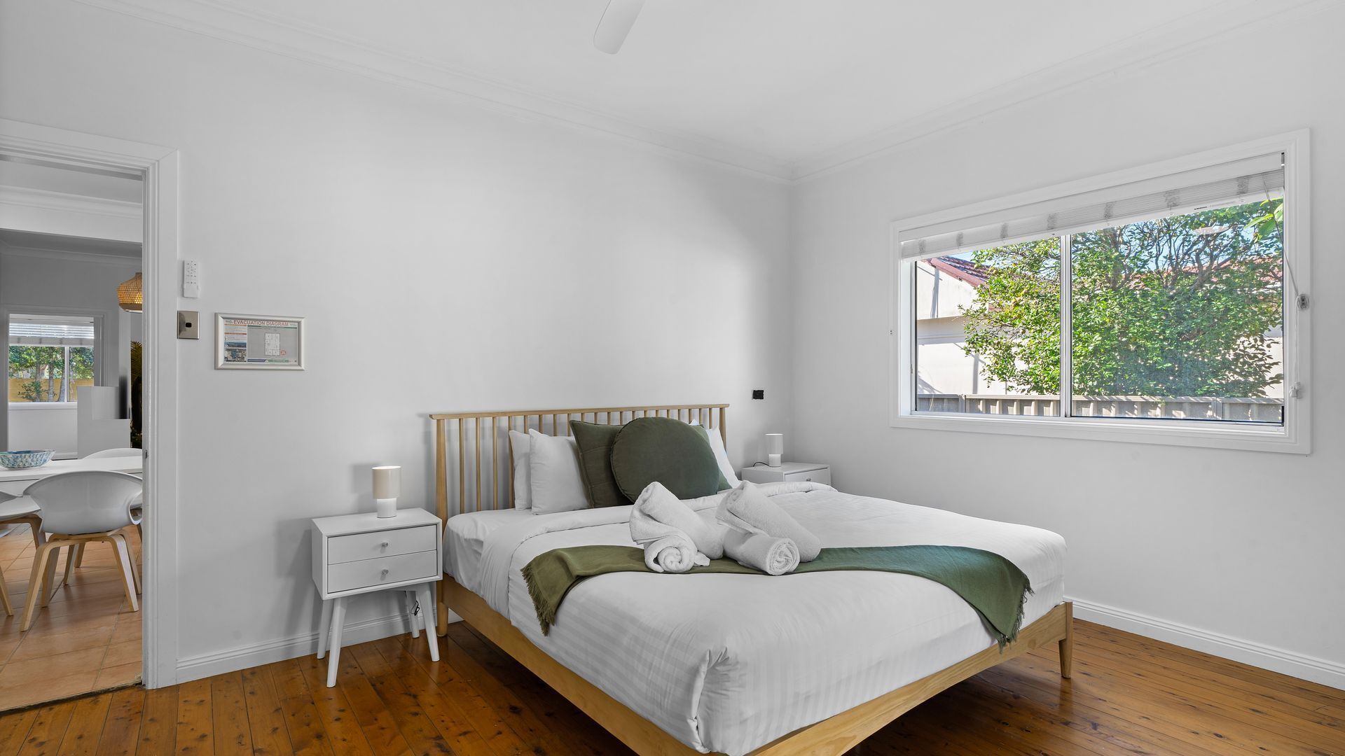 Un dormitorio con cama de madera, paredes blancas, una ventana con vistas a árboles y una mesita de noche con lámpara.