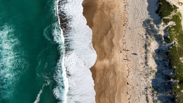 An aerial view of a beach with a sandy shore, waves crashing onto the beach, and sparse vegetation along the coastline. - The Lazy Mimosa Beach Escape Fishing, Bikes & Cozy Firepit (Budgewoi)