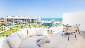 Un balcon avec un canapé blanc, deux fauteuils en osier, une petite table ornée d’une plante et de deux verres, et une vue sur un bâtiment d’un complexe hôtelier et sur l’océan.