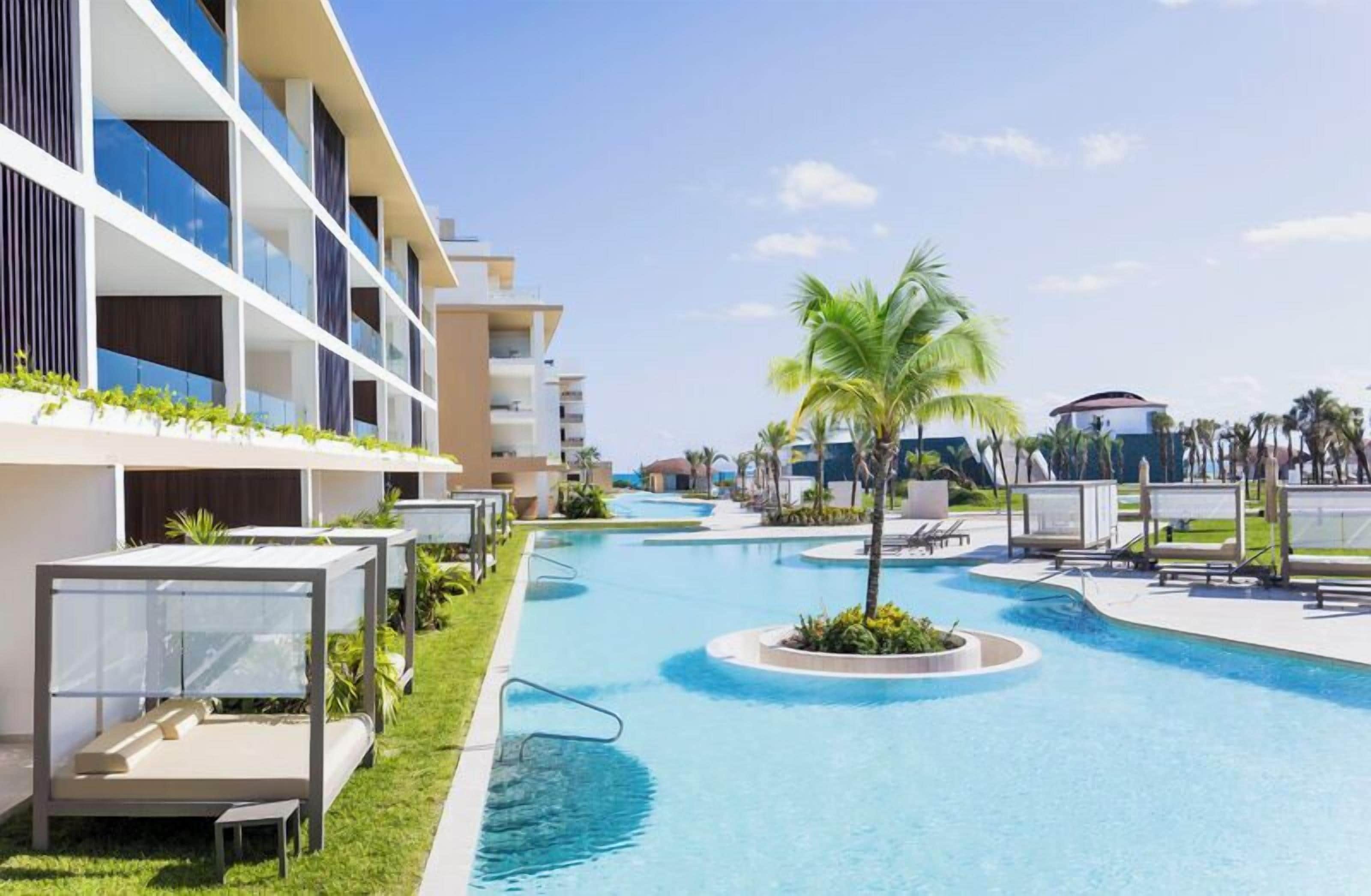 Una piscina con sillas de descanso, una palmera y un edificio de hotel con balcones.
