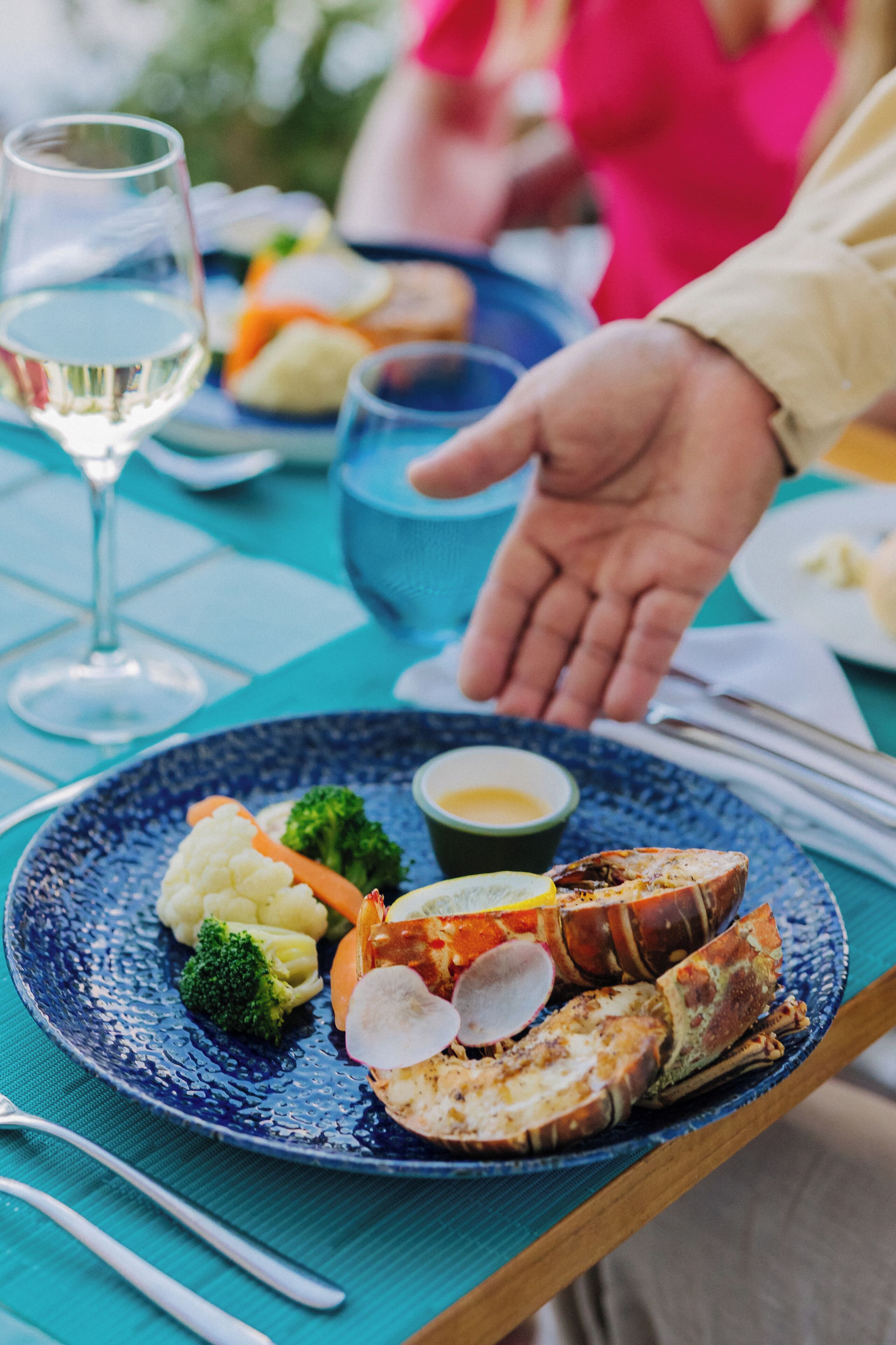 A dining table with a plate of food, a glass of white wine, and a person's hand reaching for a piece of lobster.
