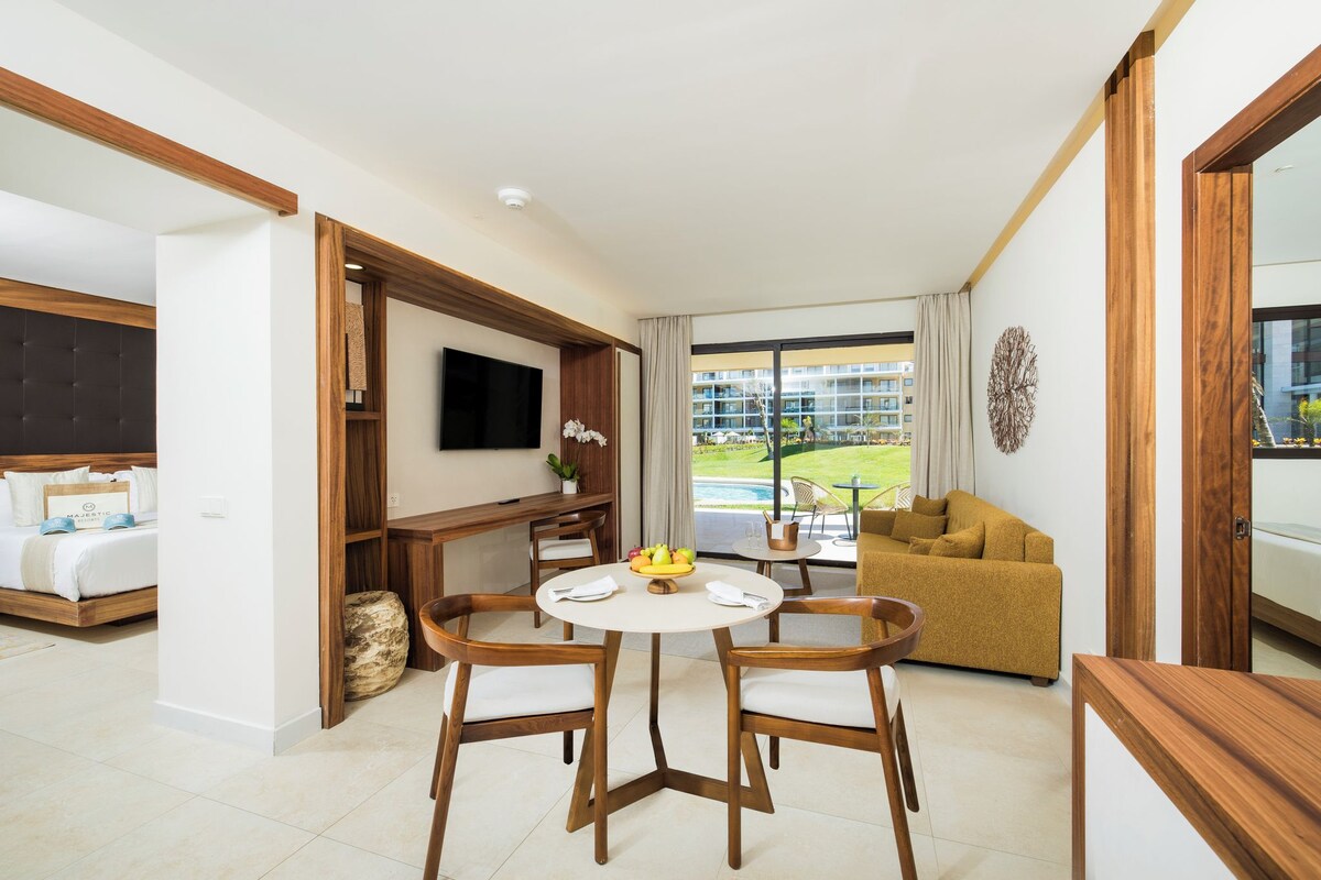 Une chambre d’hôtel moderne équipée d’un lit, d’une télévision, d’une table à manger avec des chaises, d’un canapé et offrant une vue sur une piscine et des bâtiments.