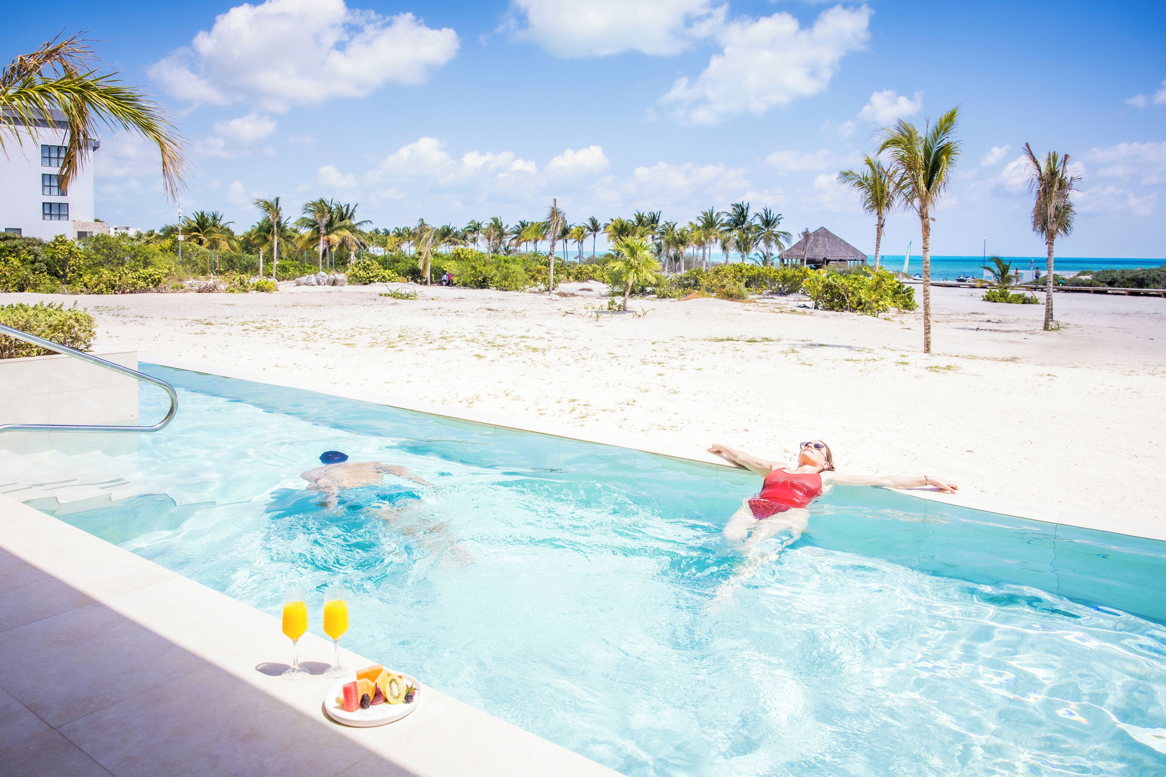 Een zwembad met twee personen in het water, een strand met palmbomen en een gebouw op de achtergrond.