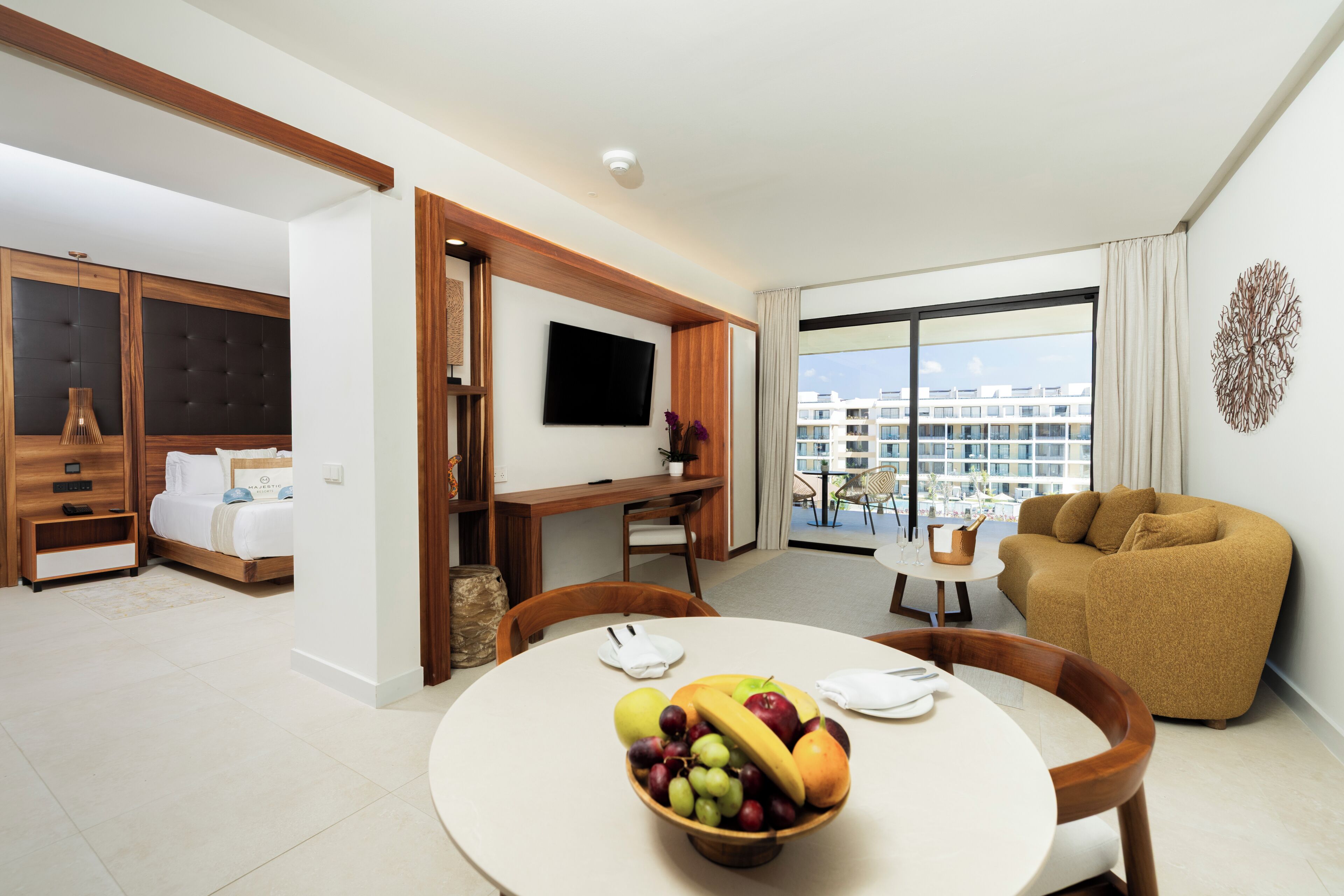 A modern hotel room with a bed, a TV, a dining area with fruit, a sofa, and a balcony with a view of buildings.