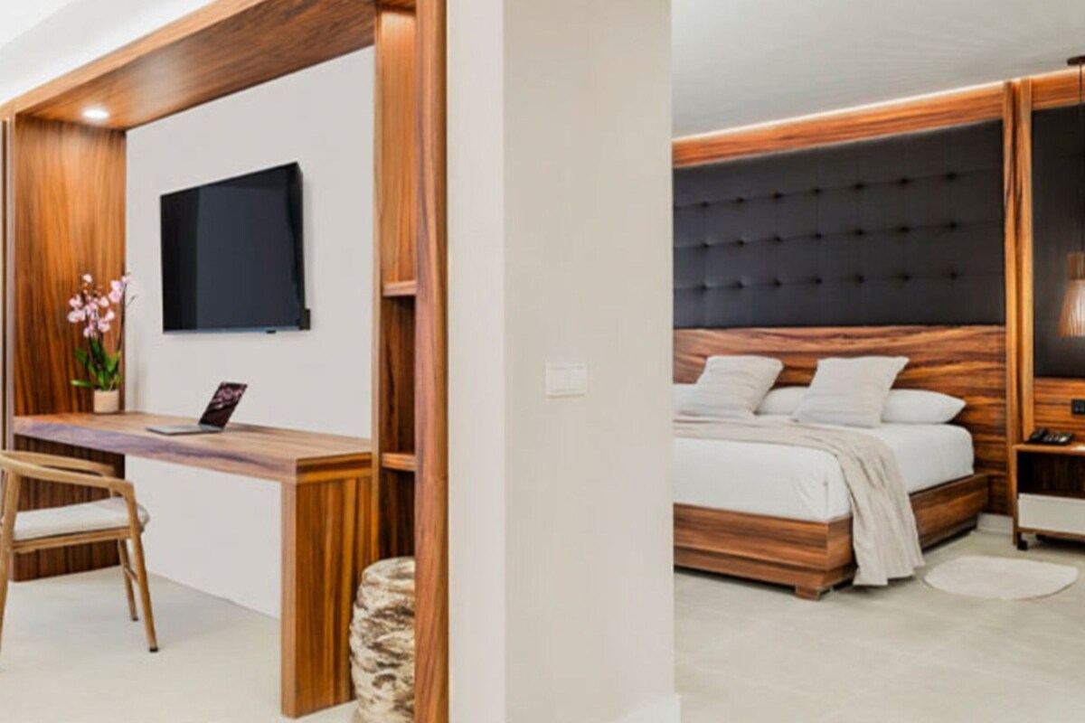 Une chambre moderne avec une tête de lit en bois, un lit recouvert de linge de lit blanc, une table de chevet et un téléviseur fixé au mur.