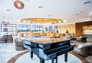Eine geräumige Lobby mit einem Flügel, bequemen Sitzgelegenheiten, großen Fenstern und einem modernen Deckenstil.