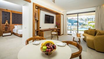 Camera d'hotel con tavolo da pranzo, divano, televisore e vista sulla piscina e sugli edifici esterni.