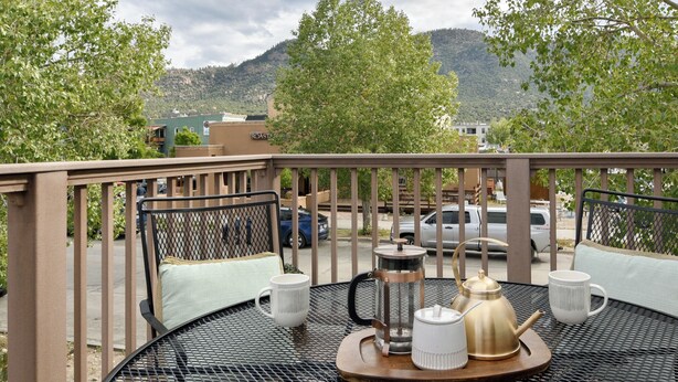 A round metal table with a tray, a coffee pot, two white cups, and a teapot on a balcony with a railing and green trees in the background.