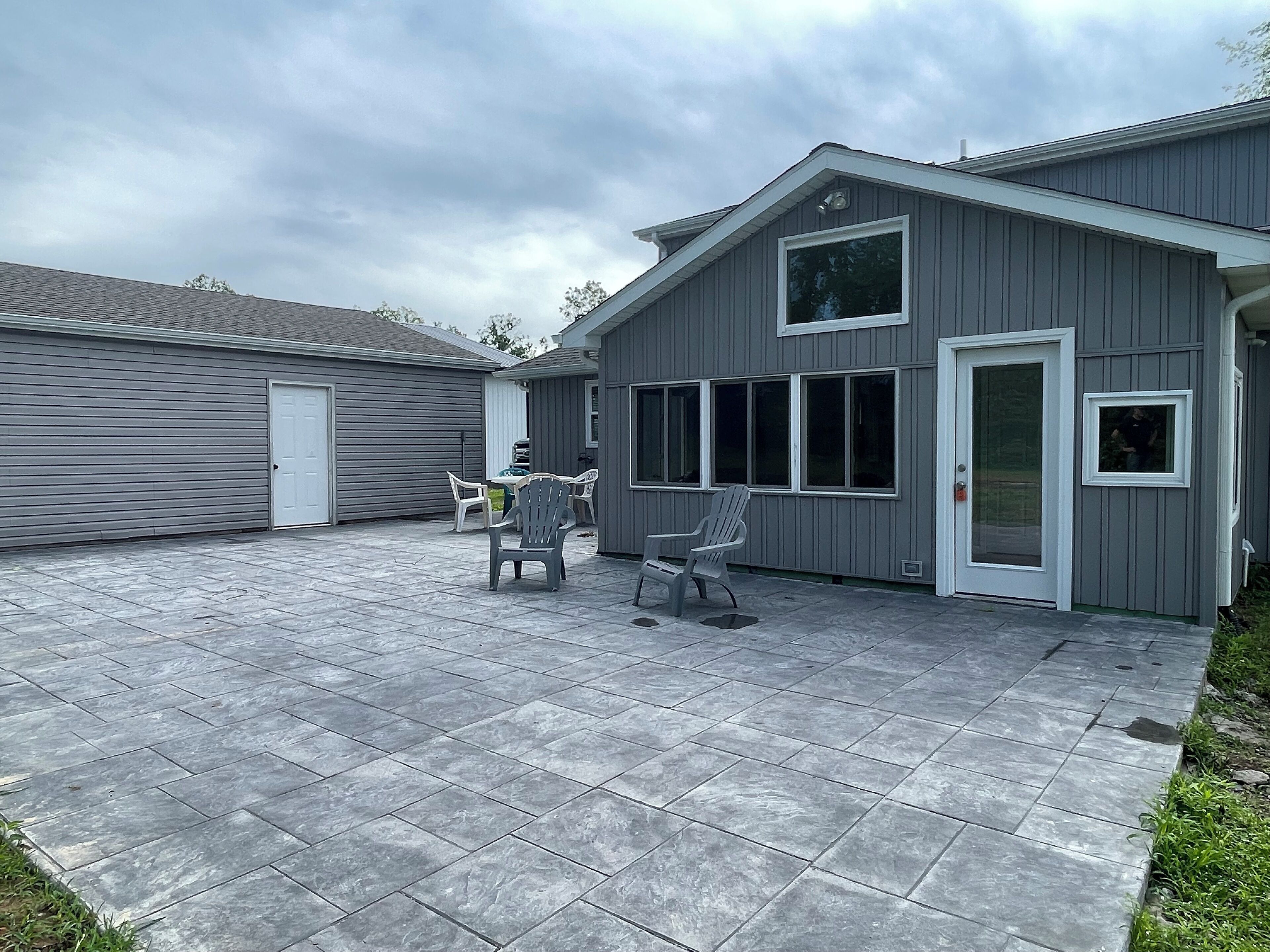 A covered patio area with grey tiles, two white plastic chairs, and a grey Adirondack chair.