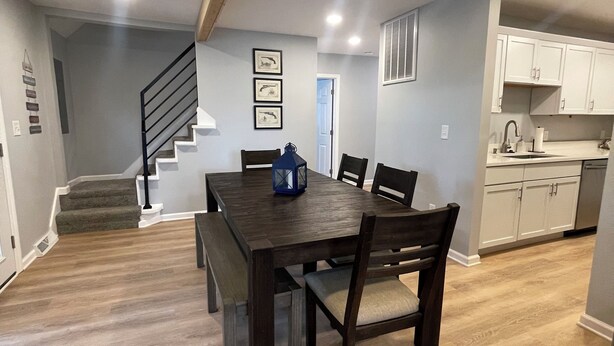 Un comedor amplio con una mesa y sillas de madera oscura, una escalera que lleva a otro nivel y una cocina con armarios blancos.