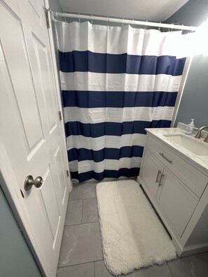 A bathroom with a white door, a white sink with a marble countertop, a white shower curtain with blue stripes, and a white fluffy rug on the floor. - Retreat to the Lake – Spacious 4BR Near The Moors Marina (Gilbertsville)