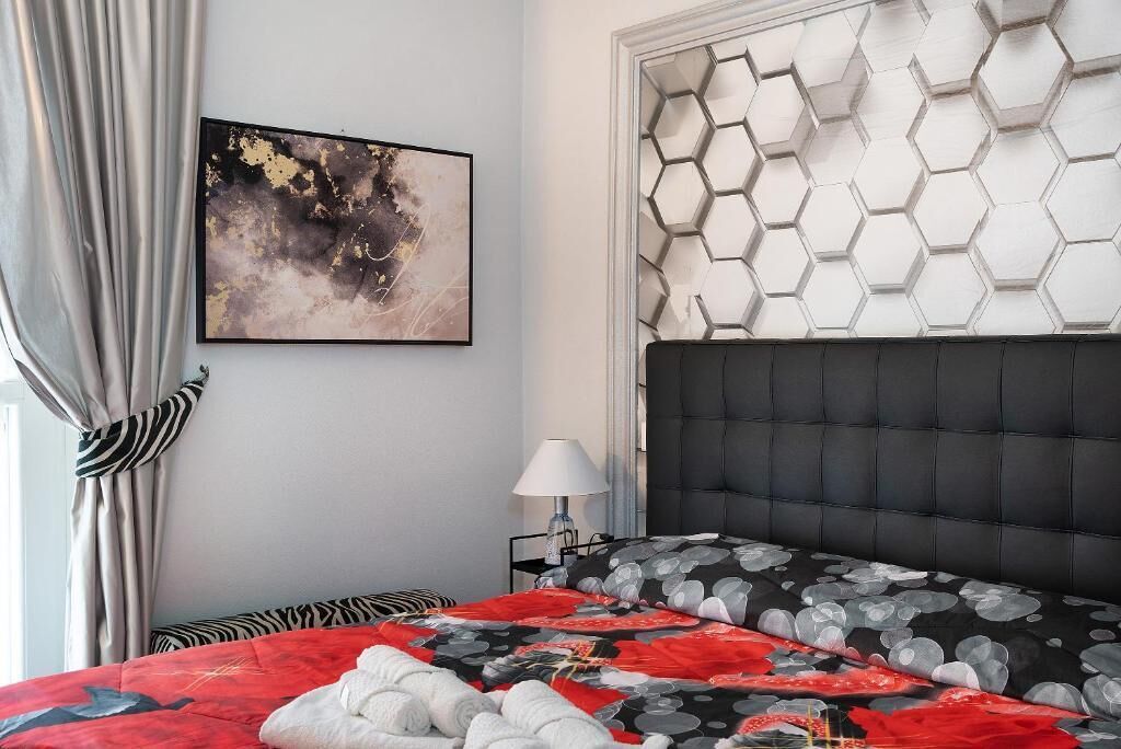 Een moderne slaapkamer met een zwart gestoffeerd hoofdeinde, een beddegoed met een rood en zwart patroon, een witte muur met een zeshoekig wandpaneel en een lijst met een abstract schilderij.